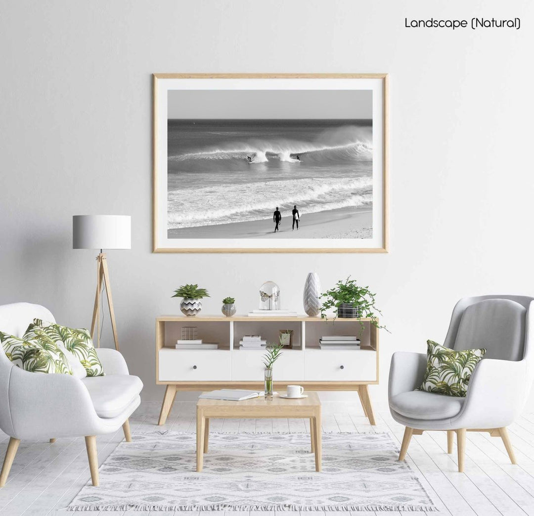 Two surfers walking on beach watching surfers on waves in black and white