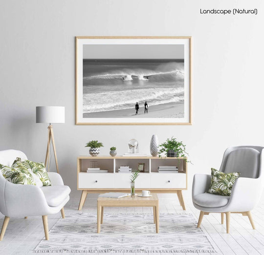 Two surfers walking on beach watching surfers on waves in black and white