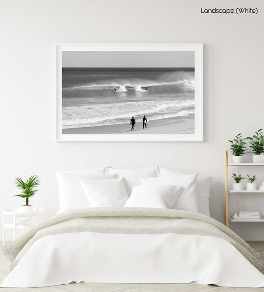 Two surfers walking on beach watching surfers on waves in black and white