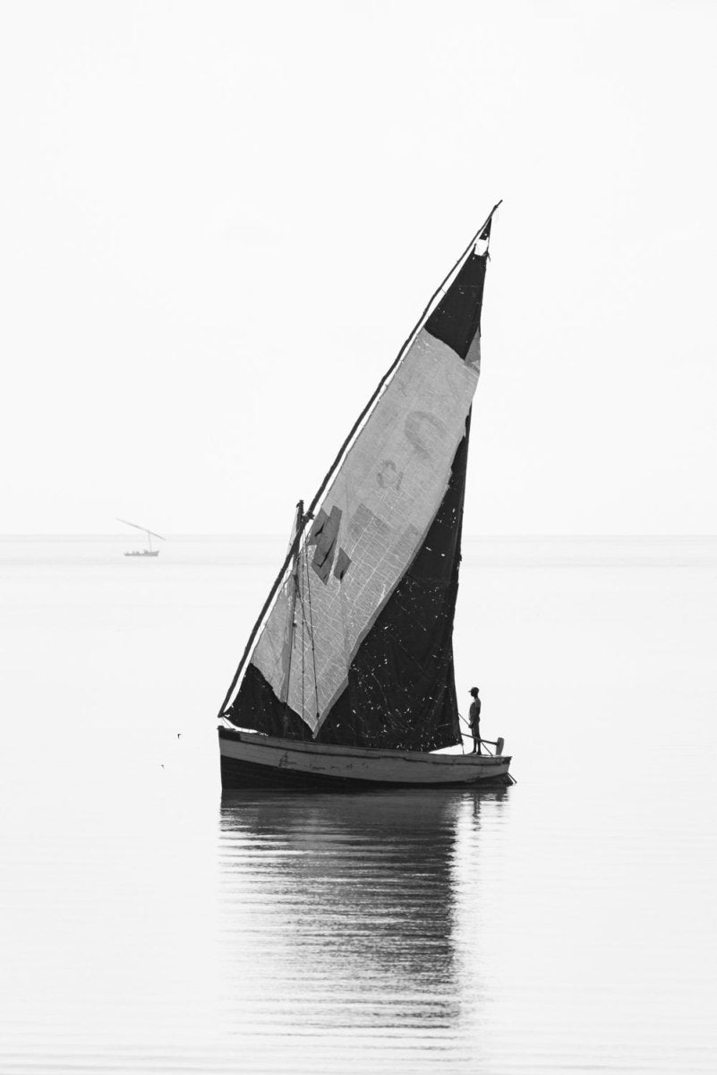 Black and white image of a single sailor on dhow boat standing tall as he anchors in the shallow calm sea in Bazaruto Archipelago, Vilankulos, Mozambique