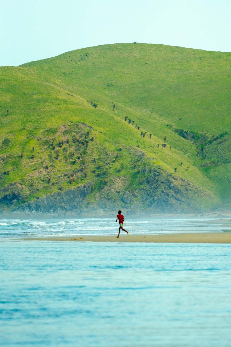 A boy runs along the beach with a large green hill as the backdrop.