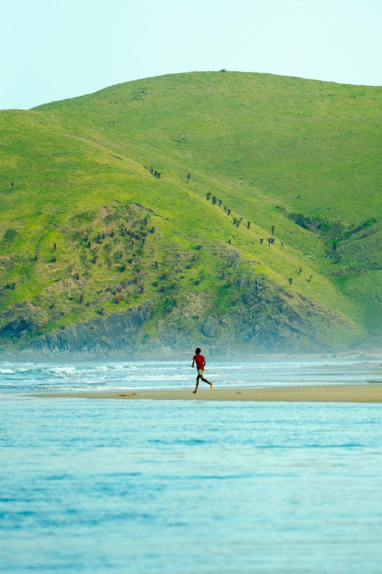 A boy runs along the beach with a large green hill as the backdrop.