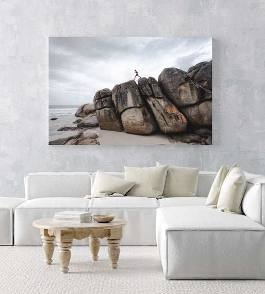 Man walking along high dark boulders on Cape Town beach in an acrylic/perspex frame