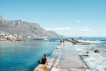 Man sitting along edge of Camps Bay pool with twelve apostles mountains in background