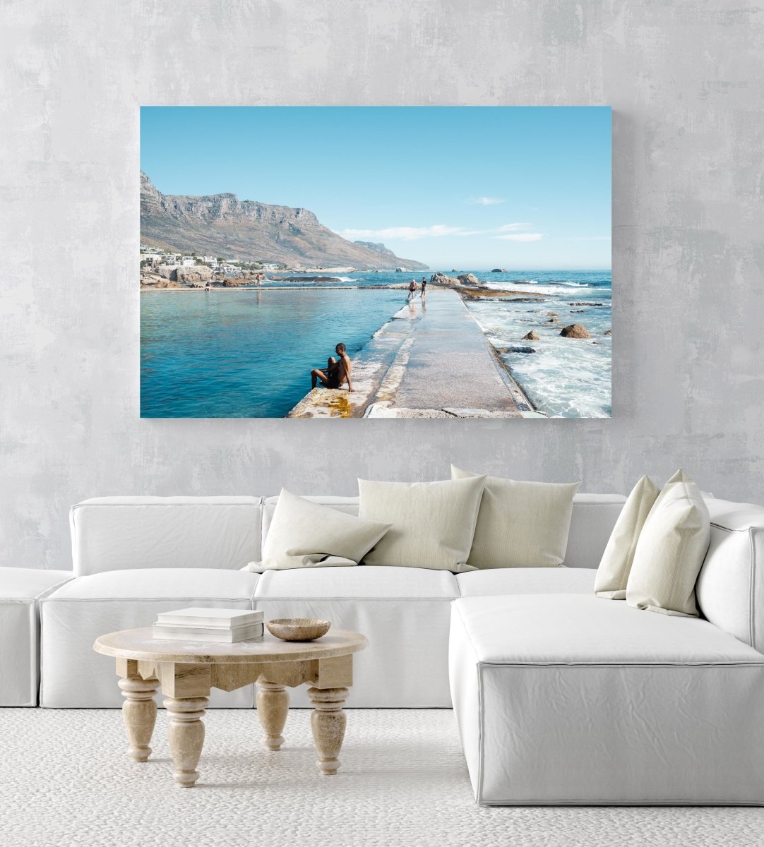 Man sitting along edge of Camps Bay pool with twelve apostles mountains in background in an acrylic/perspex frame