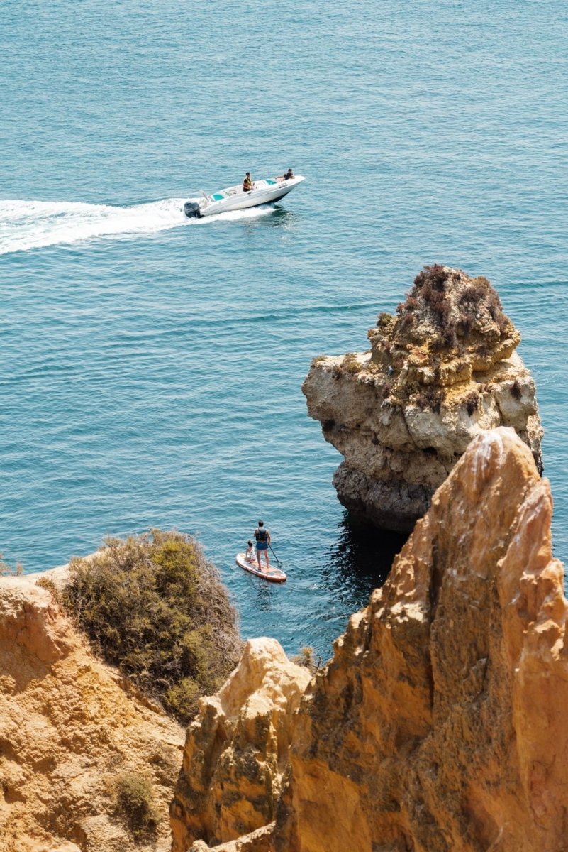 Two people on SUP watching boat drive past Lagos cliffs