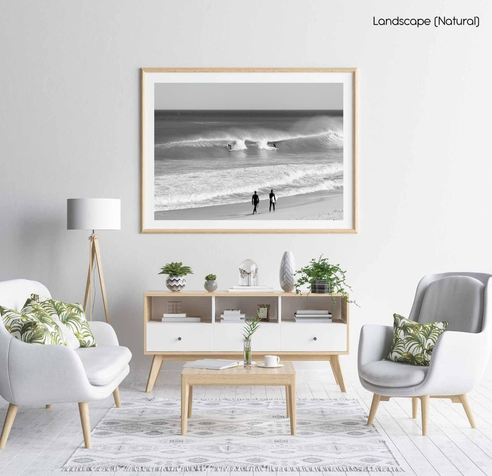 Two surfers walking on beach watching surfers on waves in black and white